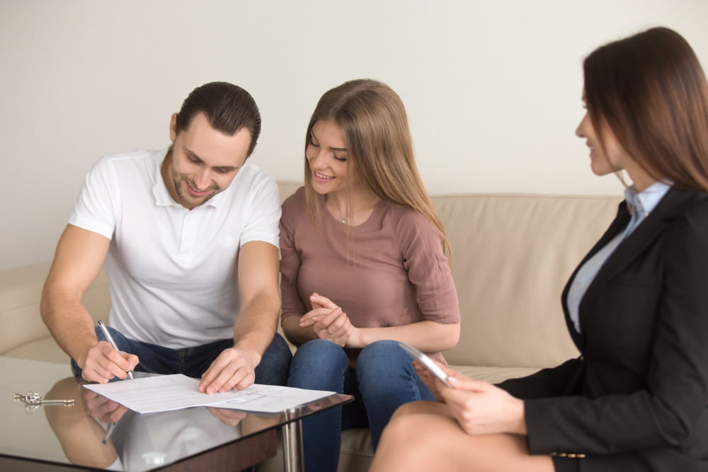 Un couple souriant assis sur un canapé en train de signer des documents officiels avec une conseillère juridique